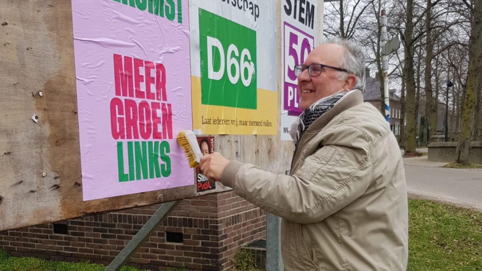 Sake plakt een GroenLinks poster op een verkiezingsbord, met een grote lach op zijn gezicht.